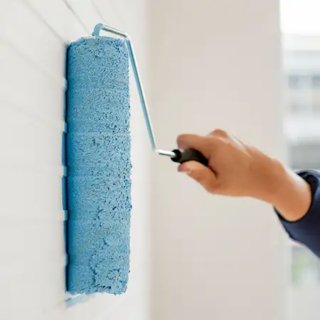 Hand streicht mit einer Farbrolle hellblaue Farbe auf eine weiße Wand.